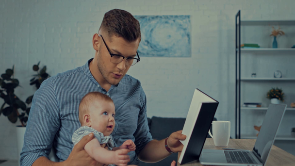 πατέρας σε γυαλιά κρατώντας το βρέφος γιο και σημειωματάριο κοντά στο φορητό υπολογιστή  - Πλάνα, βίντεο
