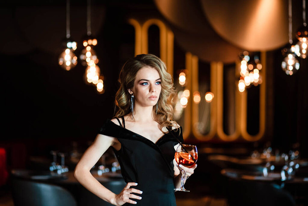 Beautiful blonde, young girl holding an aperol spritz. Cocktail aperol spritz in a glass. Portrait of a young girl in a restaurant. Portrait of a girl. Young girl in a restaurant with a glass. - Zdjęcie, obraz