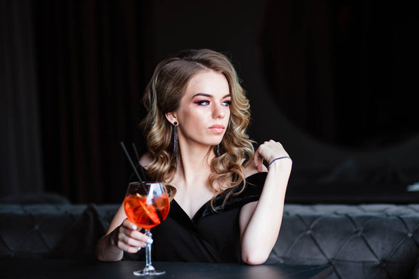Beautiful blonde, young girl holding an aperol spritz. Aperol cocktail syringe in a glass. Portrait of a young girl in a restaurant. Young girl in a restaurant with a glass on a black background. - Фото, зображення