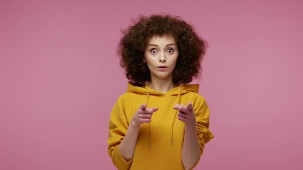 Wow, hey you! Amazed funny girl afro hairstyle in hoodie pointing finger to camera, looking big eyes surprised expression, shocked by sudden success. indoor studio shot isolated on pink background - 映像、動画