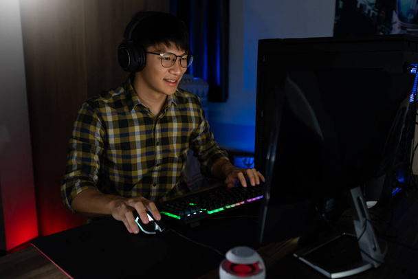 Όμορφος ενθουσιασμένος Ασιάτης gamer τύπος στα ακουστικά απολαμβάνουν και χαίροντας, ενώ παίζει βιντεοπαιχνίδια στον υπολογιστή στο άνετο δωμάτιο φωτίζεται με ζεστό και νέον φως, gaming και τεχνολογία e-sport έννοια. - Φωτογραφία, εικόνα