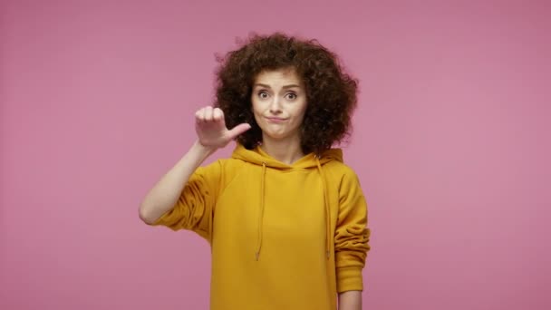 Awful result! Displeased girl afro hairstyle in hoodie showing thumbs down, dislike gesture, demonstrating disapproval disgust rejection, negative rating. studio shot isolated on pink background - Záběry, video