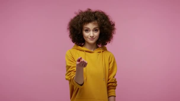 Come here, follow me! Beautiful girl afro hairstyle in hoodie making beckoning gesture with one finger, inviting to approach, looking playful flirting. indoor studio shot isolated on pink background - Imágenes, Vídeo