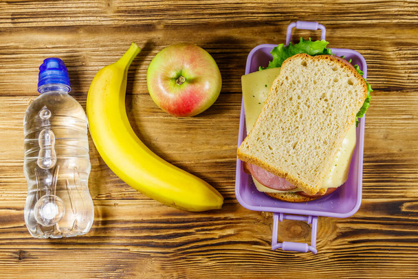 Обеденная коробка с бутербродами, бутылка воды, банан и яблоко на деревянном столе. Вид сверху - Фото, изображение