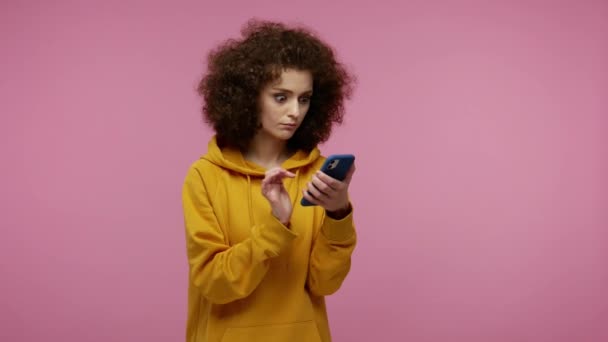 Girl afro hairstyle in hoodie scrolling social network using mobile phone and looking at camera with surprised expression, shocked by device or app. indoor studio shot isolated on pink background - Felvétel, videó