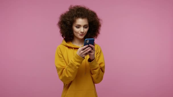 Stylish happy girl afro hairstyle in hoodie typing message on mobile phone and smiling, chatting having online communication on social network, using app. studio shot isolated on pink background - Video