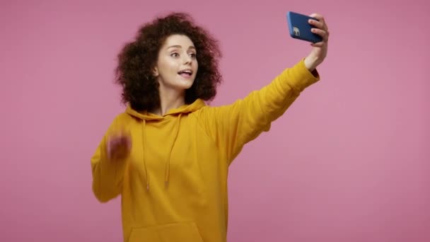 Amazing cheerful stylish girl afro hairstyle in hoodie taking selfie with positive gesture of victory, thumbs up, like, hello and sending air kiss to webcam. studio shot isolated on pink background - Imágenes, Vídeo
