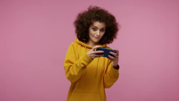 Mobile entertainment application. Girl afro hairstyle in hoodie eagerly playing video game on mobile phone, enjoying gameplay in shooter or races. indoor studio shot isolated on red background - Filmagem, Vídeo
