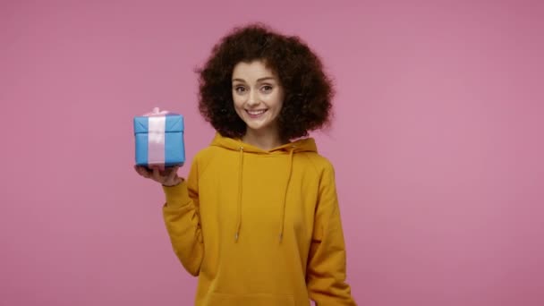 This present for you! Charming happy girl afro hairstyle in hoodie pointing at wrapped gift box and smiling, congratulating on holidays, offering bonus. indoor studio shot isolated on pink background - Záběry, video