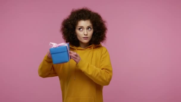 Adorable girl afro hairstyle in hoodie shaking wrapped gift box, holding near ear listening to guess what's inside, looking with curious confused expression. studio shot isolated on pink background - Imágenes, Vídeo