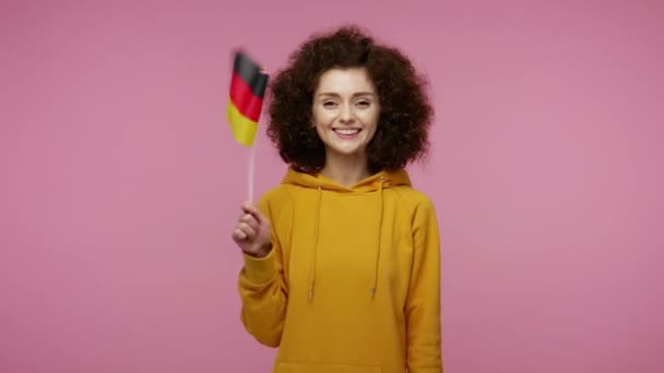 Cheerful patriotic girl afro hairstyle in hoodie waving German flag and smiling, celebrating Independence Day national holiday, human rights, democracy. indoor studio shot isolated on pink background - Кадри, відео