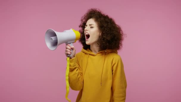 Funny girl afro hairstyle in hoodie talking with megaphone, proclaiming news, loudly announcing advertisement, warning using loudspeaker to shout speech. studio shot isolated on pink background - Imágenes, Vídeo