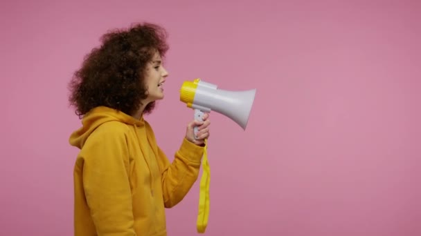 Side view, girl afro hairstyle in hoodie talking with megaphone, proclaiming news, loudly announcing advertisement, warning using loudspeaker to shout speech. studio shot isolated on pink background - Materiaali, video