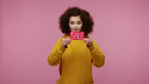 Amazed girl afro hairstyle in hoodie holding Sale word, looking at camera with surprised expression, shocked by purchase, unbelievable discounts on Black Friday. studio shot isolated, pink background - Footage, Video