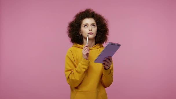 Thoughtful creative girl afro hairstyle in hoodie thinking over future business plan, taking notes in paper notebook, writing creative idea and smiling inspired happy. indoor studio shot isolated - Imágenes, Vídeo