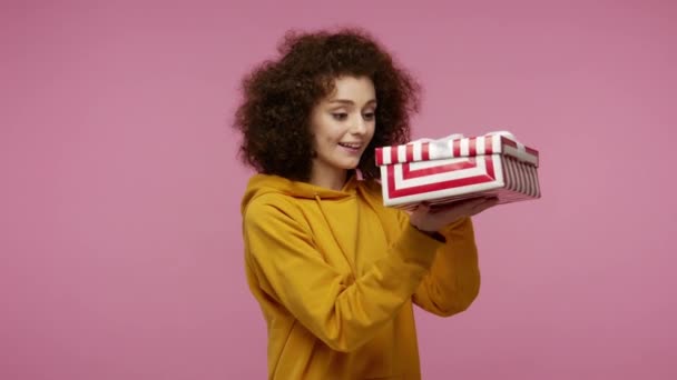 Positive girl afro hairstyle in hoodie opening gift box, unwrapping birthday surprise and expressing great happiness, showing thumbs up, satisfied with best present bonus. indoor studio shot isolated - Felvétel, videó