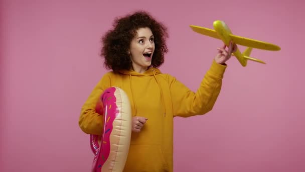 Excited tourist girl afro hairstyle in hoodie with rubber ring playing with toy airplane, dreaming of trip abroad, summer travel, vacation on seaside. indoor studio shot isolated on pink background - Кадры, видео