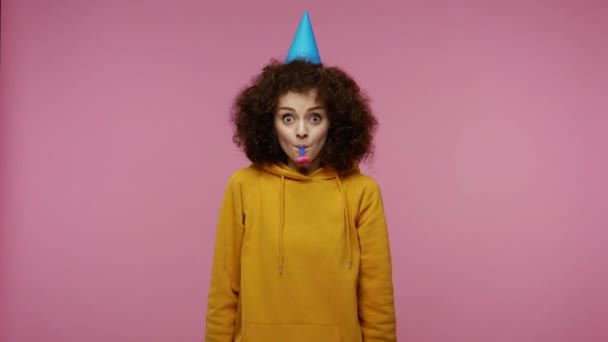 Optimistic delighted girl afro hairstyle in hoodie with funny cone on her head blowing party horn, celebrating birthday congratulating on anniversary, festive mood. indoor studio shot, pink background - Imágenes, Vídeo