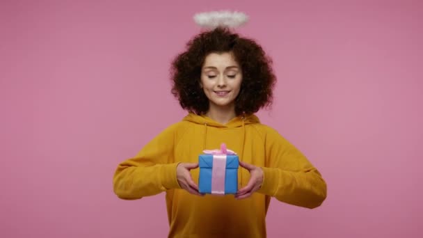 Take this present! Charming angelic girl afro hairstyle with saint halo giving wrapped gift box to camera and smiling, congratulating on holidays, donation charity concept. studio shot isolated - Záběry, video