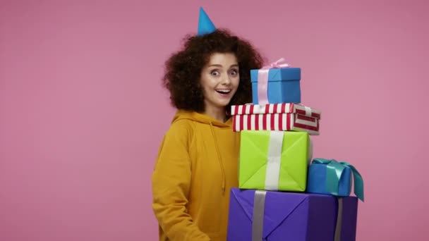 Delighted joyful girl afro hairstyle with funny cone hat holding mount of boxes, smiling excitedly satisfied with best birthday gifts, lot of presents. indoor studio shot isolated on pink background - Πλάνα, βίντεο