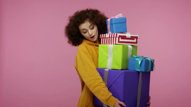 彼女の手の中にボックスのマウントでは、プレゼントの多くは、多くの休日の贈り物に興奮を見てパーカーで楽しい驚きの女の子アフロヘアスタイル。ピンクの背景に隔離された屋内スタジオで - 映像、動画