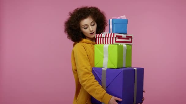 Amazed astonished girl afro hairstyle in hoodie looking surprised at mount of boxes in her hands, shocked by unbelievable birthday gifts, lot of presents. studio shot isolated on pink background - 映像、動画