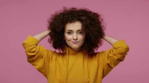 Funny girl urban style outfit putting on hood, hiding fluffy curly hair, afro hairdo, dancing and laughing, fooling around, feeling happy carefree. indoor studio shot isolated on pink background - Filmagem, Vídeo