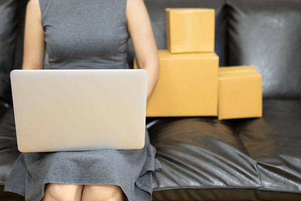 Young woman working laptop computer from home on sofa start small business in a cardboard box at work.The seller prepares the delivery box for the customer, online sales, or ecommerce.Selling online ideas concept. - Photo, Image