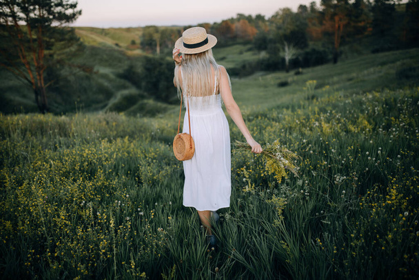 Elinde kır çiçekleri tutan şapkalı genç sarışın kız. Kadınlar güzel doğayla bir tarlada yürür. - Fotoğraf, Görsel