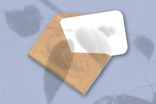 An envelope with a sheet of textured white paper on the blue background. Mockup with an overlay of plant shadows. Natural light casts shadows from an exotic plant. Horizontal orientation - Photo, Image