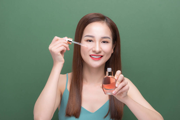 Beautiful Asian woman applying cosmetic skin serum treatment isolated on green background. - Photo, Image