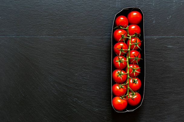 Red cherry tomatoes in a paper try placed vertically on black background with copy space. Natural and organic vegetables full of vitamins and nutrients for a healthy lifestyle - Photo, Image