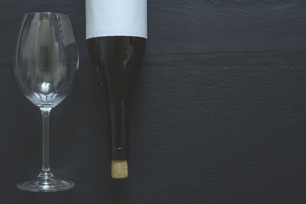 Σκούρο μπουκάλι κόκκινο κρασί με λευκή ετικέτα και μεγάλο κομψό ποτήρι κρασιού σε μαύρο τραπέζι με χώρο αντίγραφο. Γλυκό ποτό σερβίρεται σε φανταχτερά εστιατόρια ως έρημο - Φωτογραφία, εικόνα