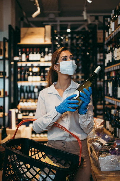 Beautiful young and elegant woman with face protective mask and gloves buying healthy food and drink in a modern supermarket or grocery store. Pandemic or epidemic lifestyle and consumerism concept. - Φωτογραφία, εικόνα