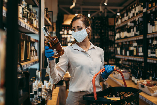 Beautiful young and elegant woman with face protective mask and gloves buying healthy food and drink in a modern supermarket or grocery store. Pandemic or epidemic lifestyle and consumerism concept. - 写真・画像