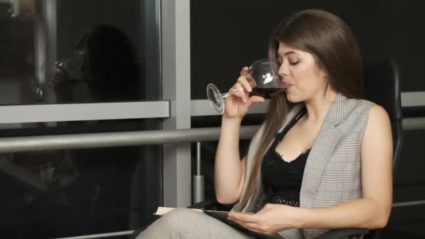 Beautiful girl of Caucasian appearance with long hair sitting by the panoramic window reads a book holding a glass of red wine and enjoys the night city - Metraje, vídeo