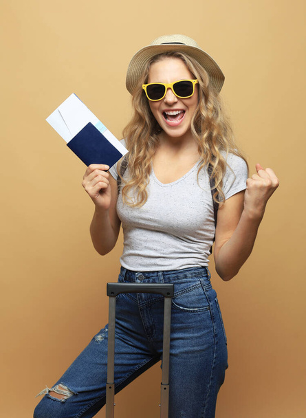 Cheerful blonde woman in sunglasses posing with baggage and holding passport with tickets over beige background - Valokuva, kuva