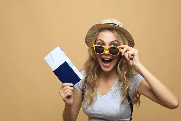 Beautiful blonde woman in sunglasses posing with passport with tickets over beige background - Foto, afbeelding