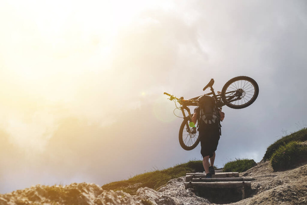 homem esporte em ação levantamento de bicicleta em seu ombro na montanha de rocha com fundo céu nublado
. - Foto, Imagem