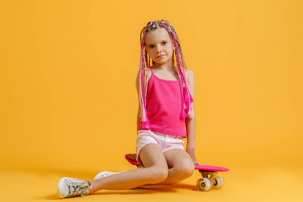 Active young girl in pink shirt and shorts sitting on skateboard over yellow background - Фото, зображення