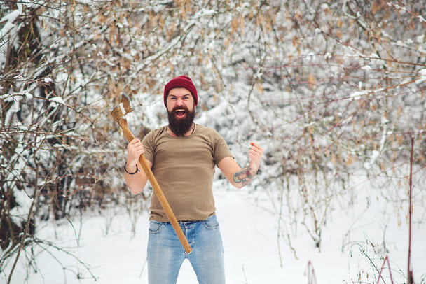 Karlı ormanda sakallı oduncu. Kış parkında baltası olan kaba sakallı bir adam. Acımasız sakallı adam olumsuz duygular gösteriyor. Hippi adam kışın dışarıda baltasını tutuyor.. - Fotoğraf, Görsel