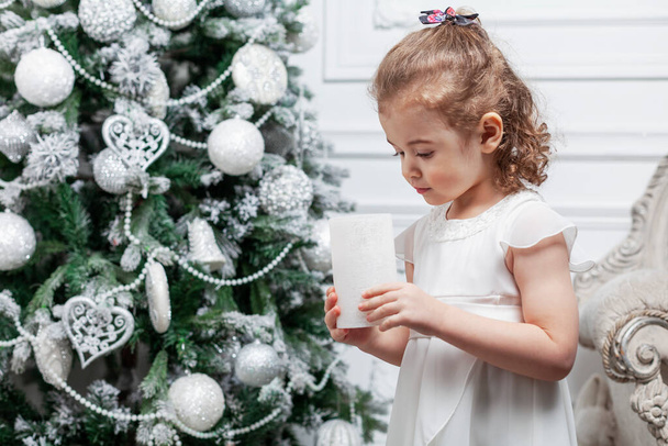 A little girl in a beautiful white dress stands near a toy tree decorated with toys. New Year and Christmas celebration. - Foto, immagini