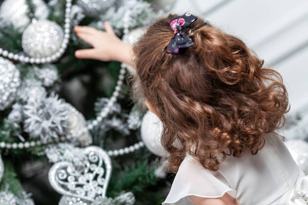 A little girl in a beautiful white dress stands near a toy tree decorated with toys. New Year and Christmas celebration. Back view. - Фото, зображення
