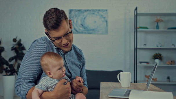 knappe vader met behulp van laptop tijdens het praten op de smartphone en het houden van baby - Video