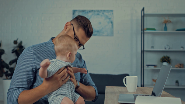 padre che punta con il dito al computer portatile mentre tiene in braccio il figlio neonato - Filmati, video