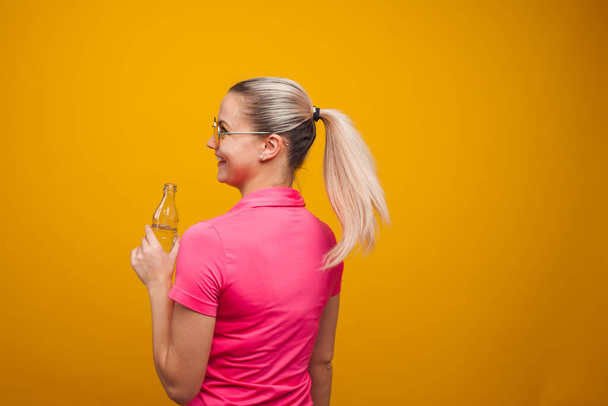 Cute blonde in sunglasses and t-shirt, drinking water from a bottle, portrait on a yellow background - Foto, Imagen