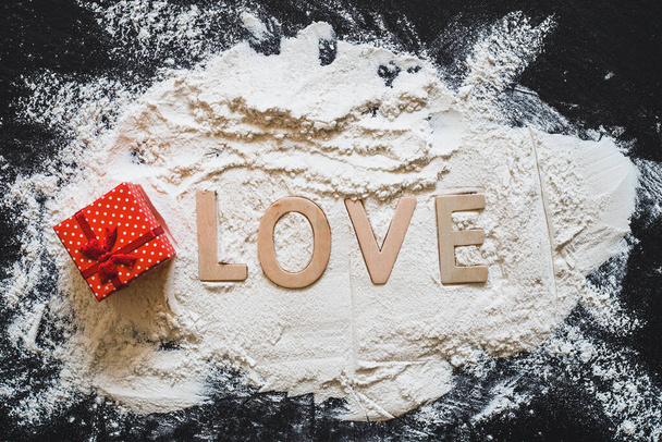 Weißes Mehl mit dem Wort Liebe und eine kleine rote Geschenkschachtel auf schwarzem Hintergrund. Vollkornpuder mit schöner Botschaft und Geschenk verteilt auf einer dunklen Steinoberfläche - Foto, Bild