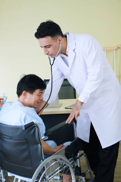 Młody kukański lekarz w mundurze w sukni Check up by Stetoskop for Asian Staruszek siedzi na wózku inwalidzkim w szpitalu. Koncepcja opieki zdrowotnej i starszego społeczeństwa - Zdjęcie, obraz