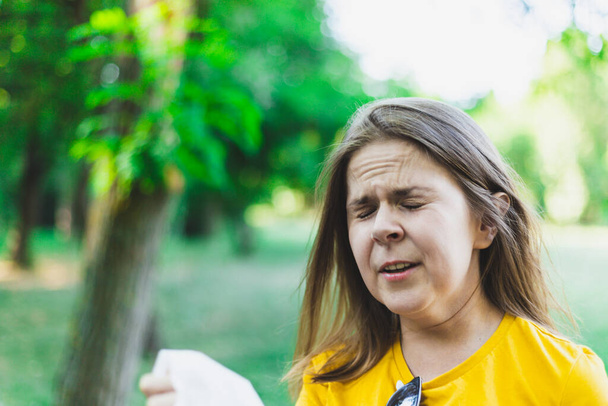 Young woman holding napkin while sneezing from seasonal allergies in the park. Beautiful girl with brown hair having flu and cold symptoms and a runny nose outdoors - Photo, Image