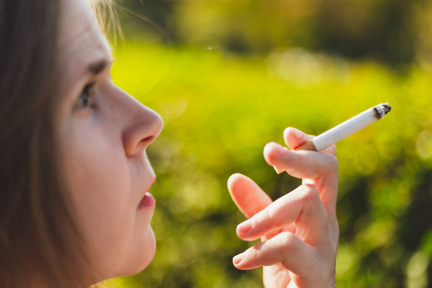 Κοντινό πλάνο με μια νεαρή γυναίκα να καπνίζει έξω. Όμορφο κορίτσι με επικίνδυνη και τοξική συνήθεια κρατώντας ένα αναμμένο πούρο στο πάρκο - Φωτογραφία, εικόνα
